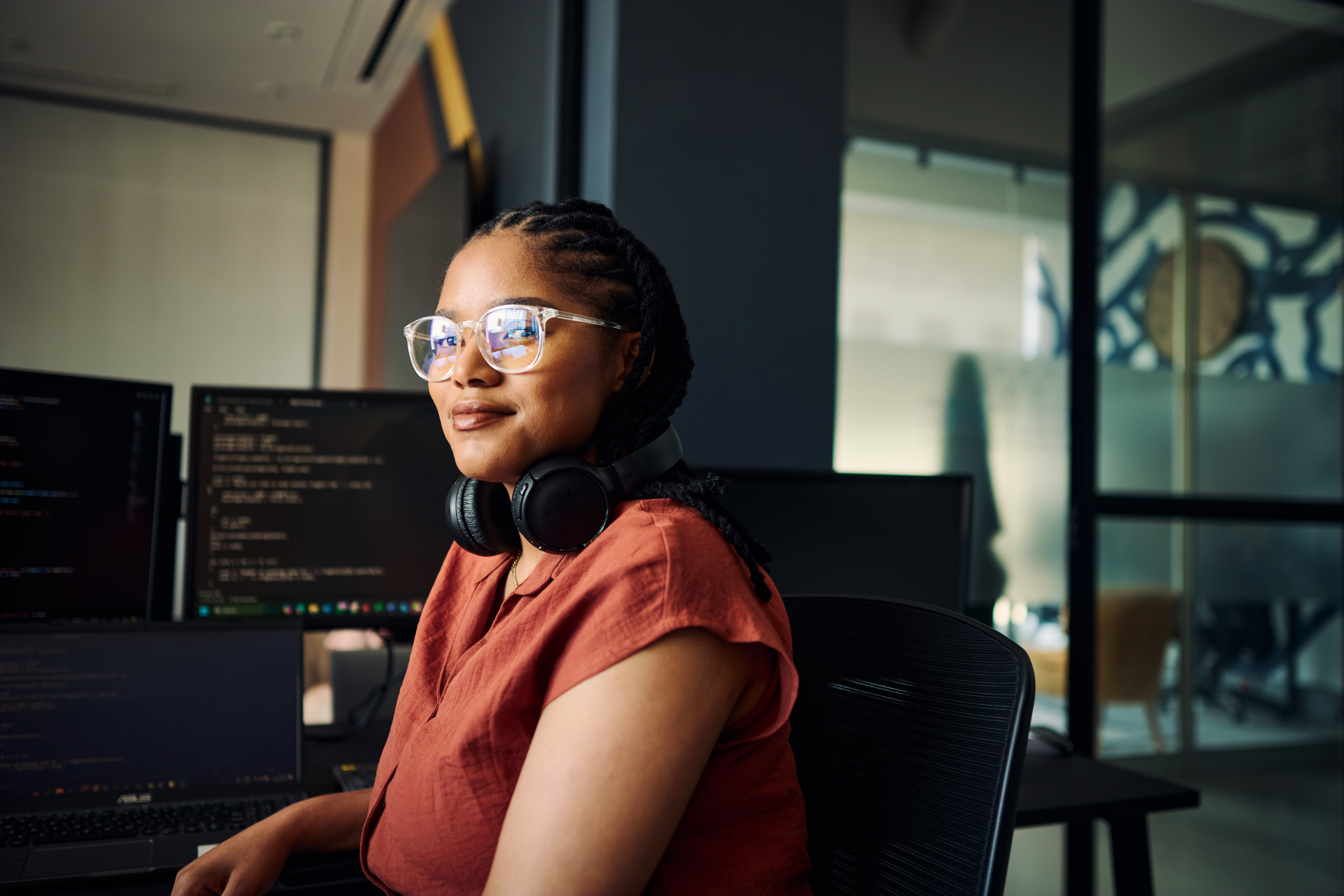 Woman developer with braids, glasses, and headphones coding at dark monitors